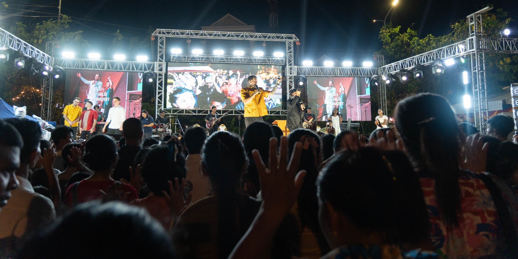 Chris preaching on the lit crusade stage as the crowd raises their hands in Kandal, Cambodia